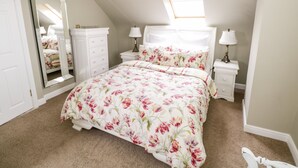 A bedroom with a sloped ceiling, a large mirror, a bed with floral bedding, two bedside tables with lamps, and a white wardrobe. - Curragh Hill (Mulranny)