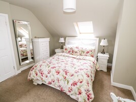 Un dormitorio con techo inclinado, un espejo grande, una cama con ropa de cama floral, dos mesitas de noche con lámparas y un armario blanco.