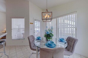 Un coin repas comprenant une table en verre, quatre chaises, un luminaire suspendu et de grandes fenêtres équipées de stores.