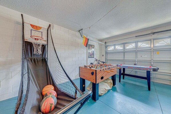 Un panier de basket, une table de baby-foot et une table de ping-pong dans une pièce avec un mur en briques et une porte de garage.