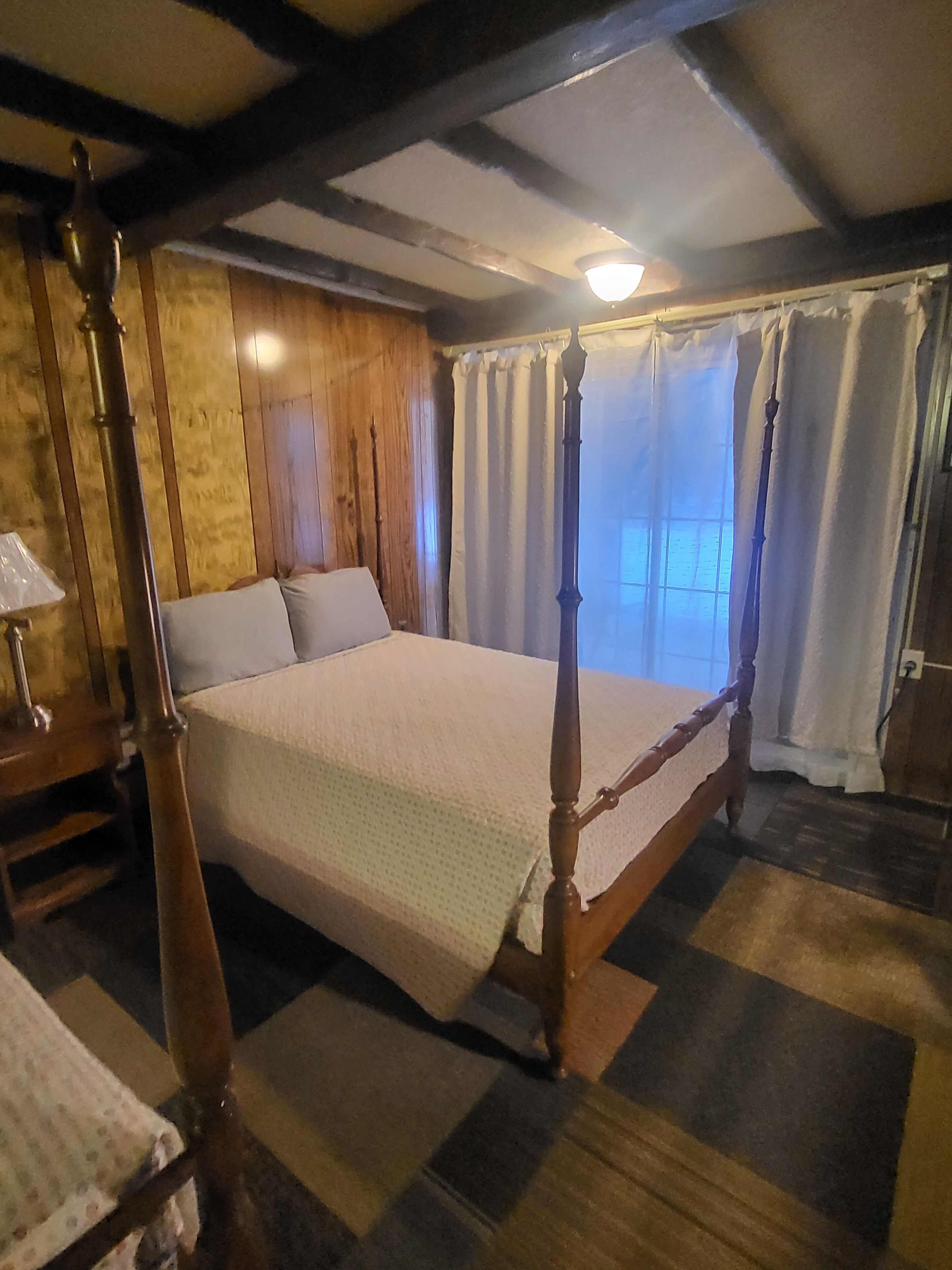 A four-poster bed with a wooden frame, a quilted bedspread, and a window with curtains.