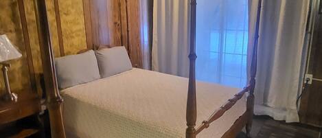 A four-poster bed with a wooden frame, a quilted bedspread, and a window with curtains.