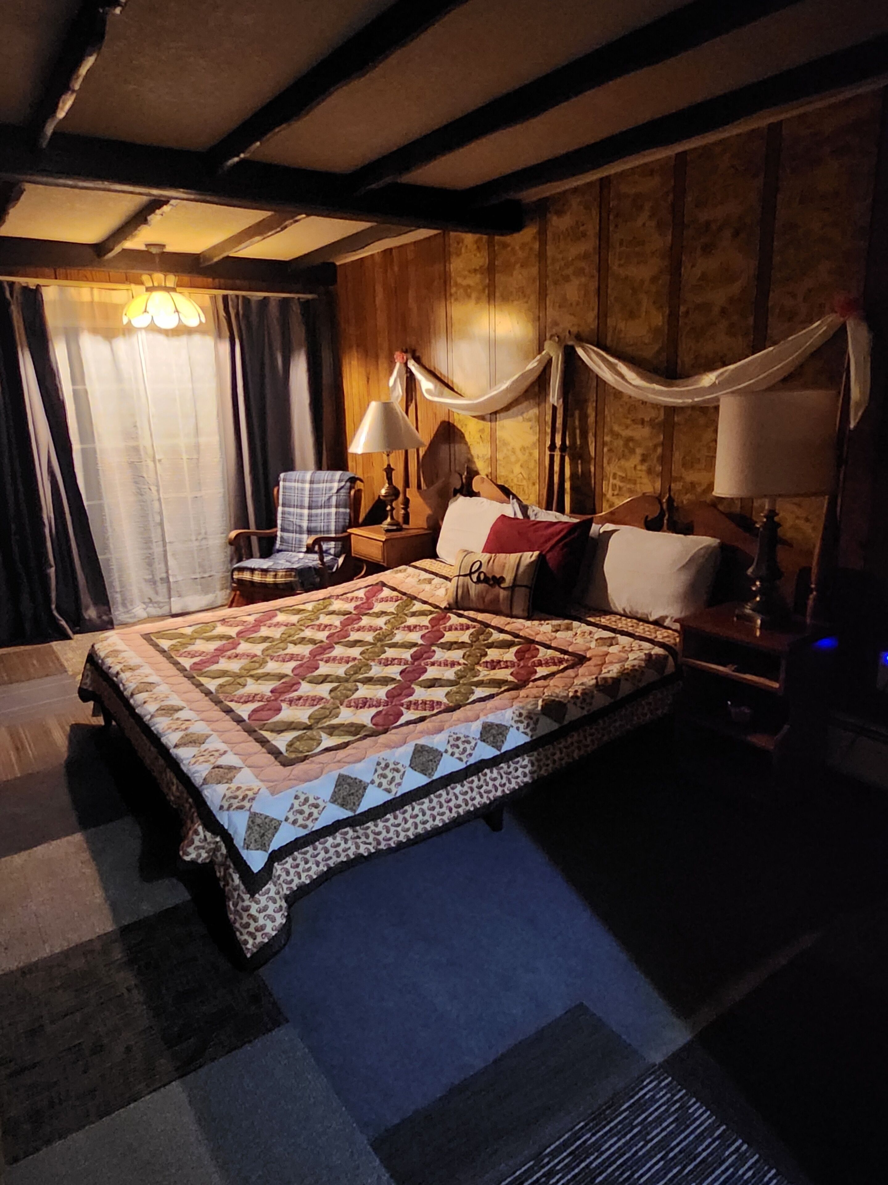 Un dormitorio con una cama grande, paredes de madera, una araña y una silla.