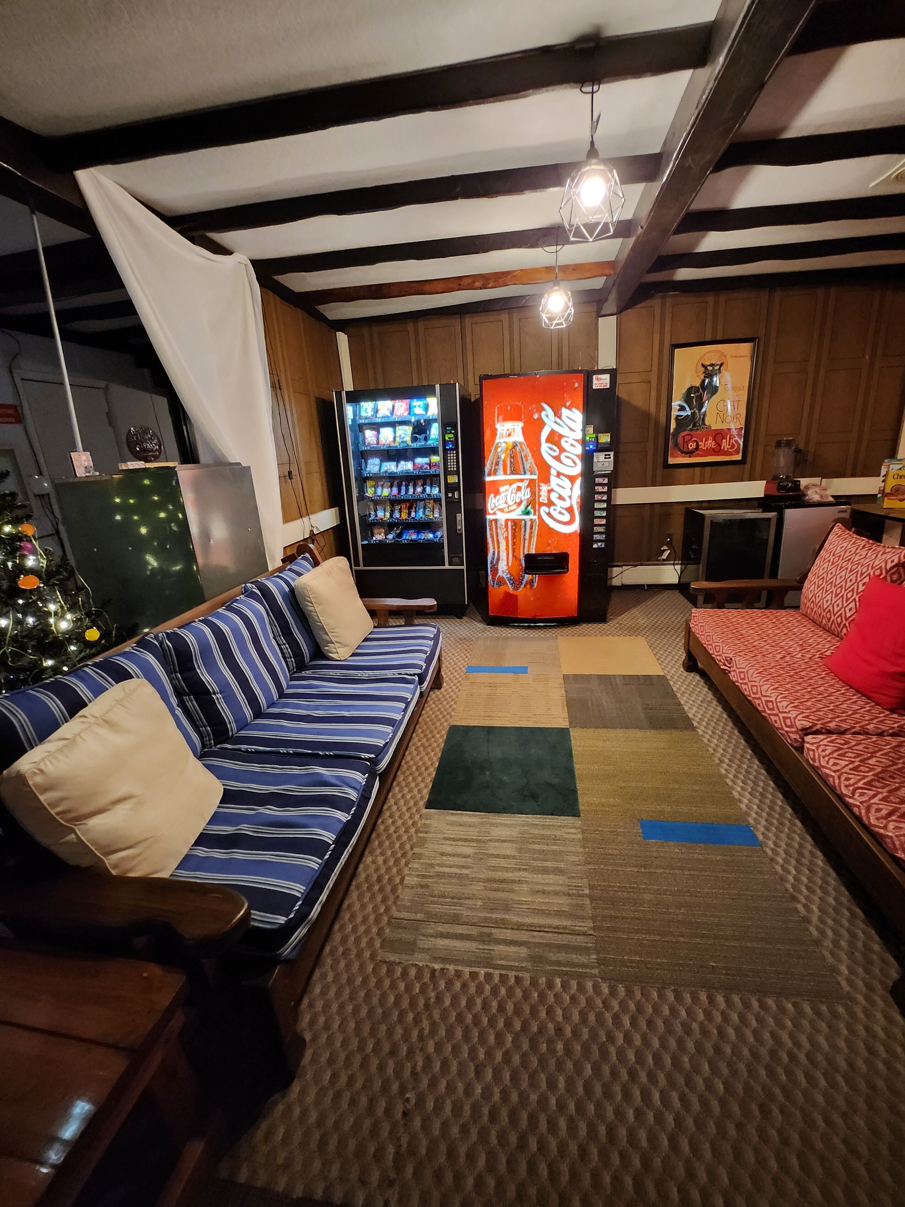 A cozy living room with a striped sofa, a red patterned sofa, and a coffee table. There are two vending machines, one with a Coca-Cola logo and another with snacks. A Christmas tree is visible in the corner.