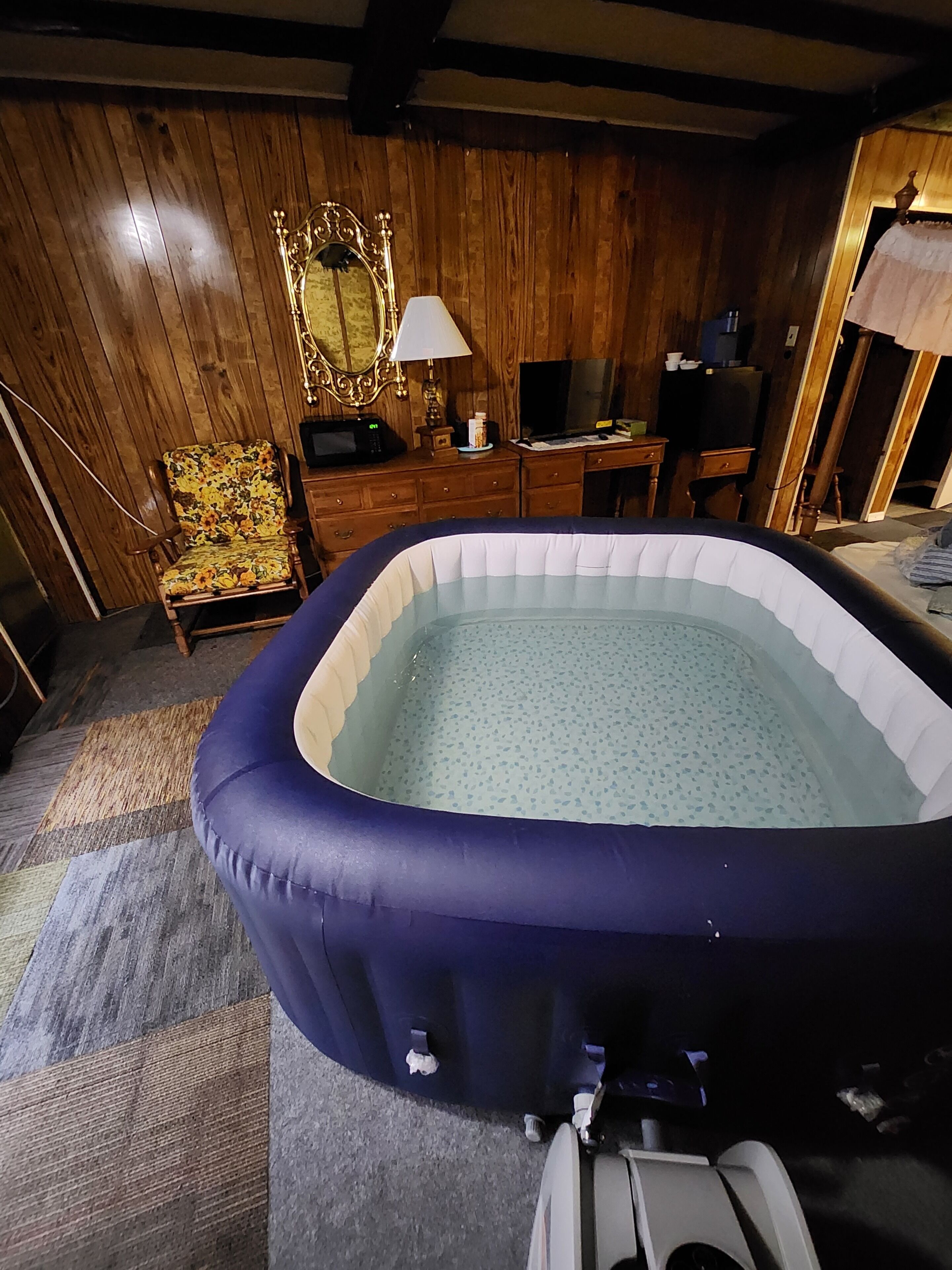 Ein Zimmer mit Holzboden, einer blauen aufblasbaren Whirlwanne, einem Holzstuhl mit floralem Sitz, einem Holztisch mit Fernseher und einem Spiegel an der Wand.