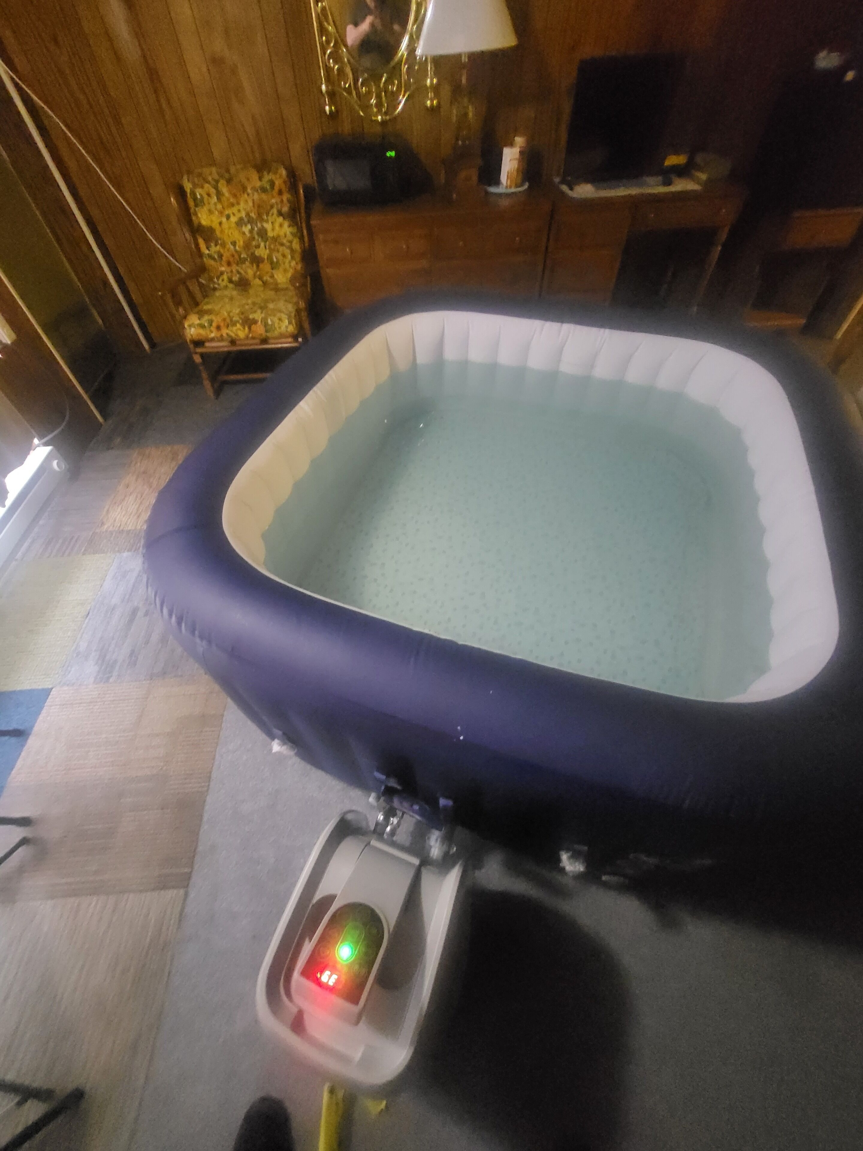 Habitación con una piscina inflable pequeña, suelo de madera, una silla y una mesa con lámpara.