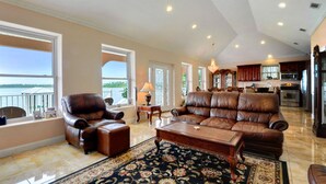 A spacious living room with a large brown leather sofa, a coffee table, and a patterned rug. There is a view of a balcony through large windows. - Sunset bay view 1 (Pass Christian)
