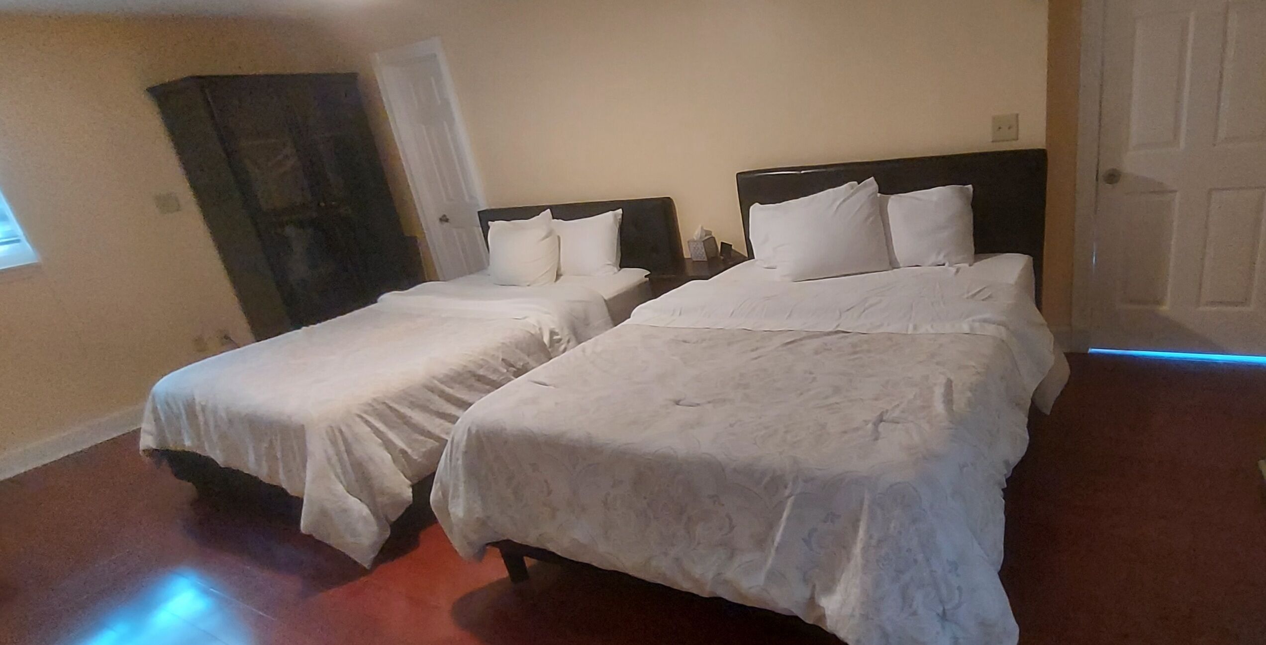 A room with two beds, each with white linens and headboards. There is a dark-colored wardrobe and a white door.