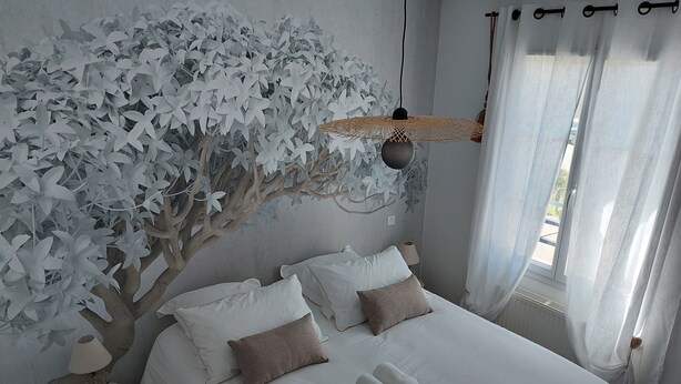 Un letto rifatto con lenzuola bianche e due asciugamani arrotolati, una lampada a parete e un albero decorativo con foglie bianche.