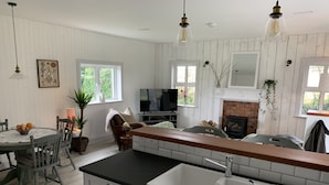 A modern kitchen with white cabinetry, a black countertop, and a built-in sink. There is a dining area with a table and chairs, a fireplace, and a television. - Garden Cottage on Tintern Trails (New Ross)
