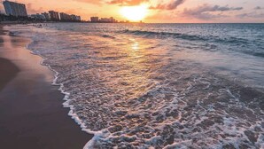 A beach with waves gently lapping onto the shore, a sunset in the background, and buildings in the distance. - OASIS 2BR/2Bath STEPS from BEACH + Parking @ ESJ (Carolina)