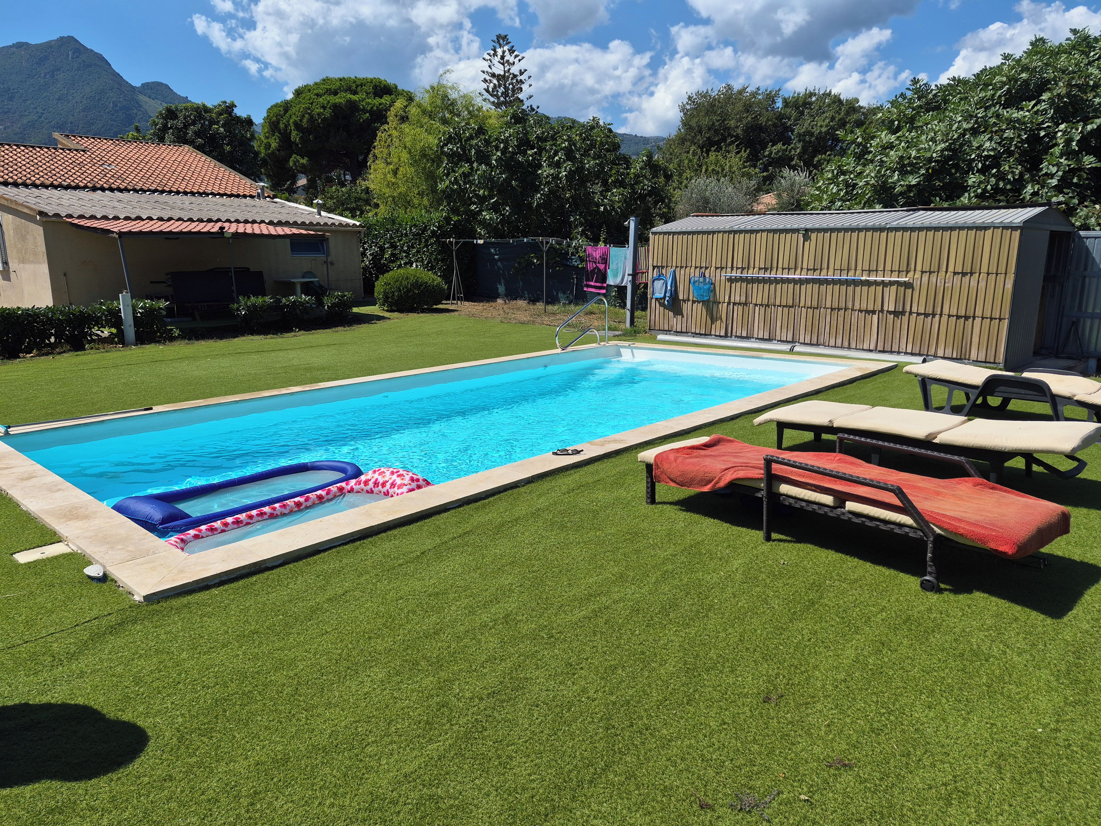 Une piscine avec un toboggan, entourée de gazon synthétique, de chaises longues et d’un abri en bois.