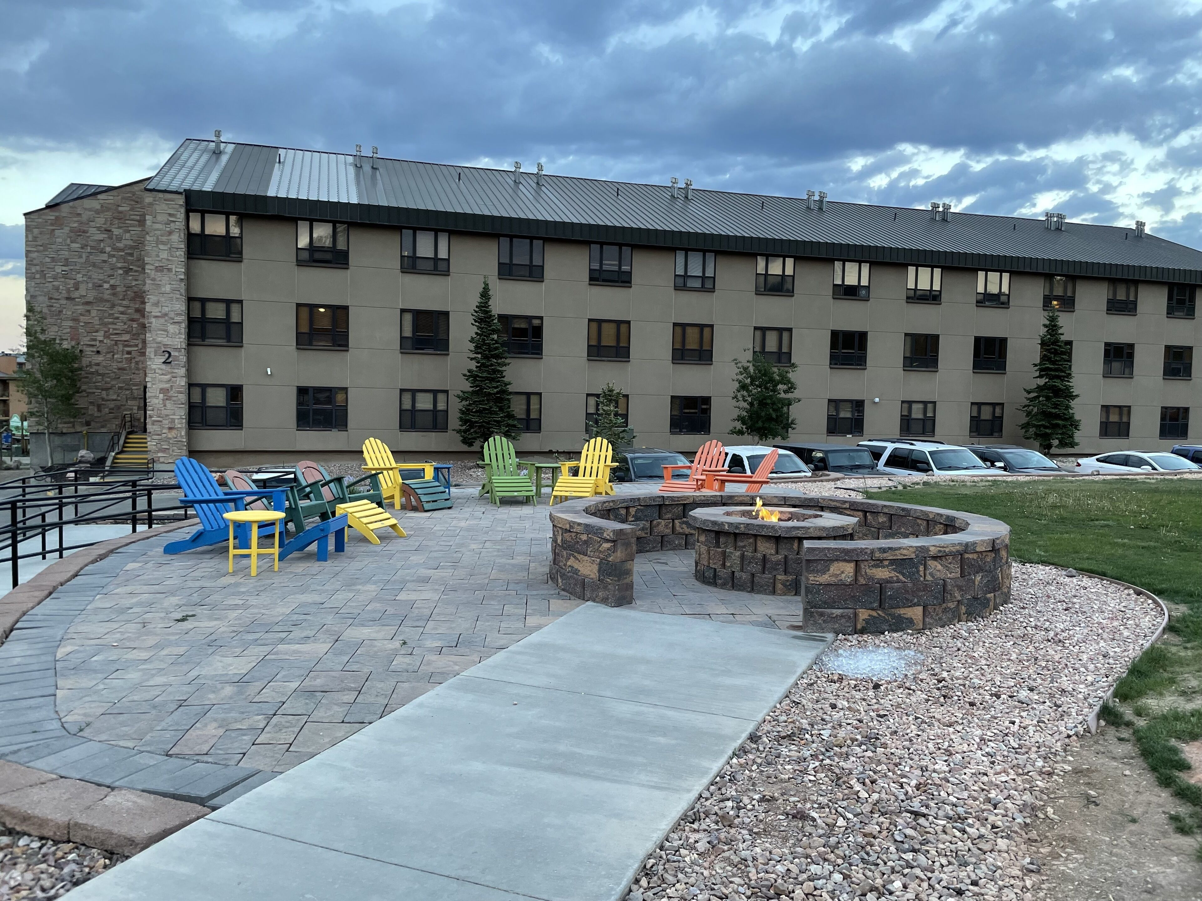 A building with a stone facade and multiple windows. There is a paved patio area with colorful Adirondack chairs and a fire pit.