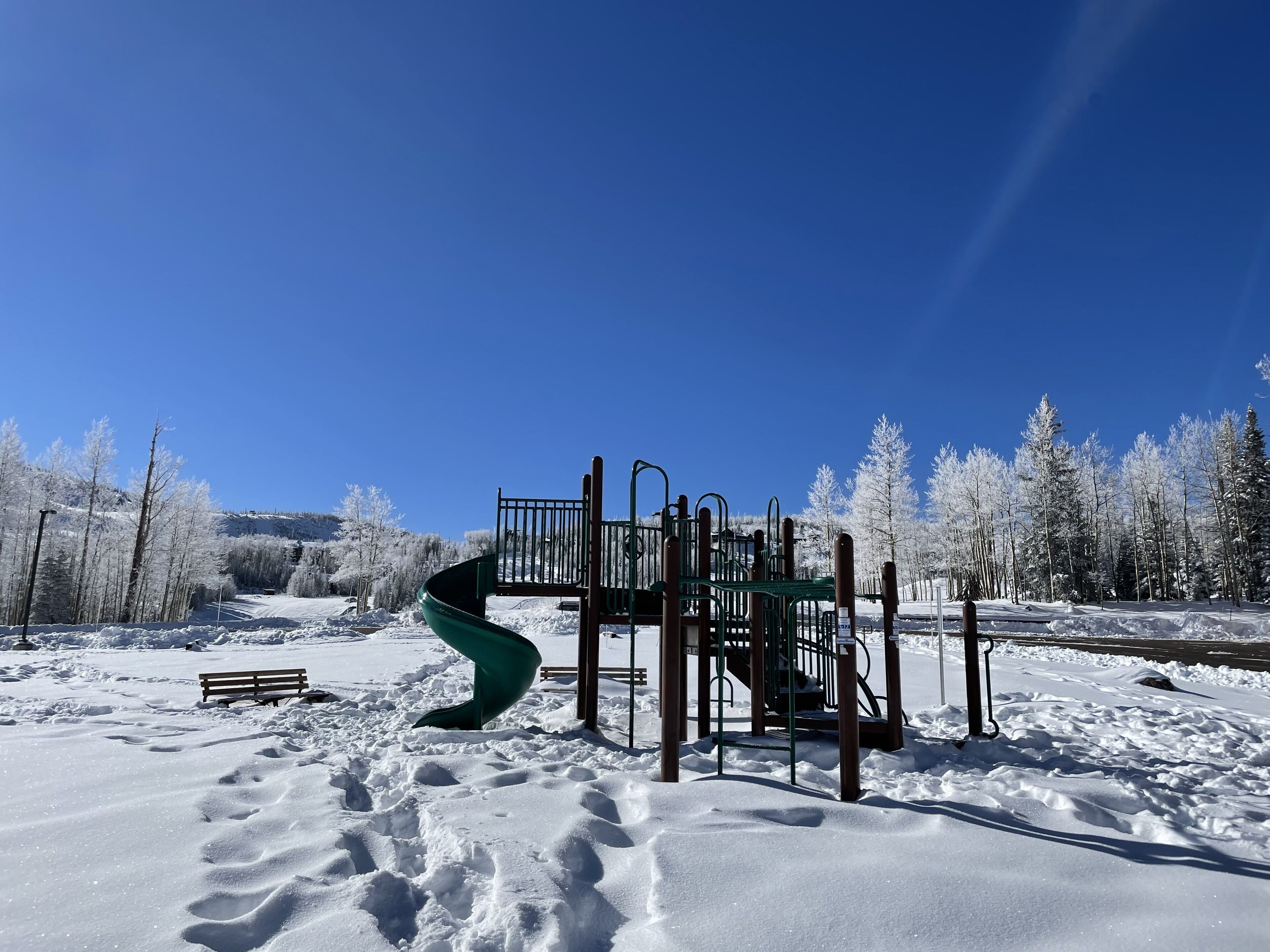 Een speeltuin met een spiraalvormige glijbaan en klimconstructies, omgeven door met sneeuw bedekte bomen en een helderblauwe lucht.