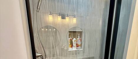 Una ducha sin barreras con puerta de vidrio, pared de mármol y un estante con artículos de aseo.