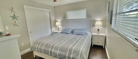 Une chambre à coucher comprenant un lit, deux tables de chevet avec des lampes, un ventilateur de plafond, une fenêtre avec des stores et une porte.