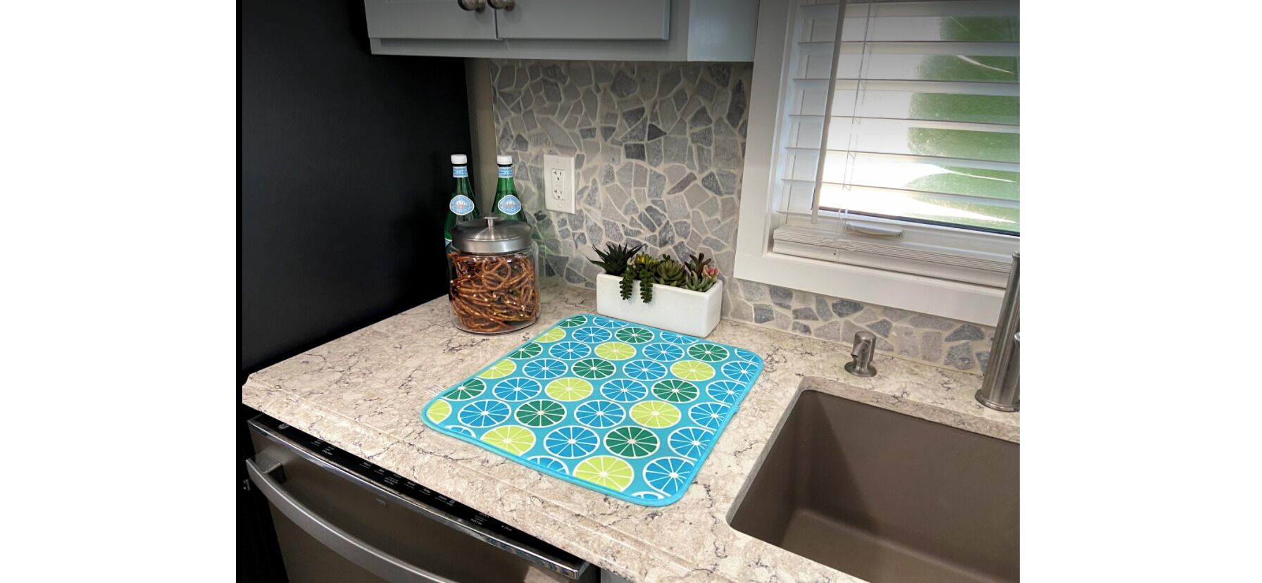 A kitchen with a granite countertop, a blue and green citrus-patterned mat, a jar of pretzels, and a small potted plant.