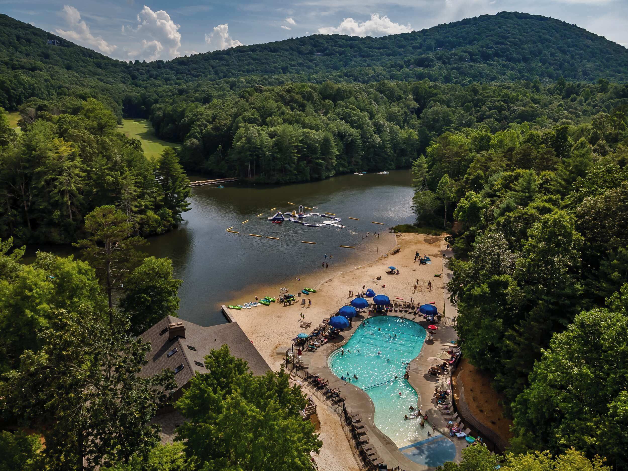 A resort with a swimming pool, beach, and boats on a lake surrounded by forested hills.