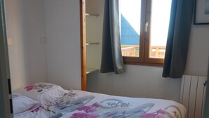 A single bed with a floral bedspread, a wooden floor, a window with curtains, and a wall-mounted light fixture. - Appart "les luges" 4/6 people valley view large balcony swimming pool  (Besse-et-Saint-Anastaise)