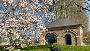A brick building with an arched entrance, surrounded by a grassy area with scattered white petals, and a large flowering tree in the background. - Little cottage (Thiouville)