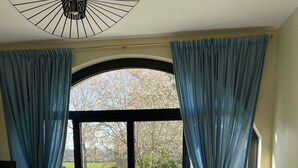 A modern living room with a large sliding glass door leading to a garden. There is a sofa, a dining table with chairs, and a lamp. - Little cottage (Thiouville)