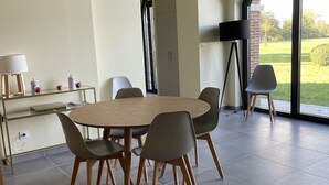 A modern dining area with a round table and six chairs, a small side table with a lamp, and a view of a garden through large windows. - Little cottage (Thiouville)