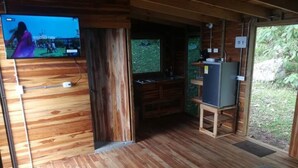 L’intérieur d’un chalet en bois, avec un téléviseur fixé au mur, un petit coin cuisine et une vue sur l’extérieur.