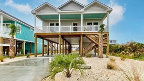 A beachfront house with a covered porch, a staircase leading to a second level, and a sandy beach in the foreground.