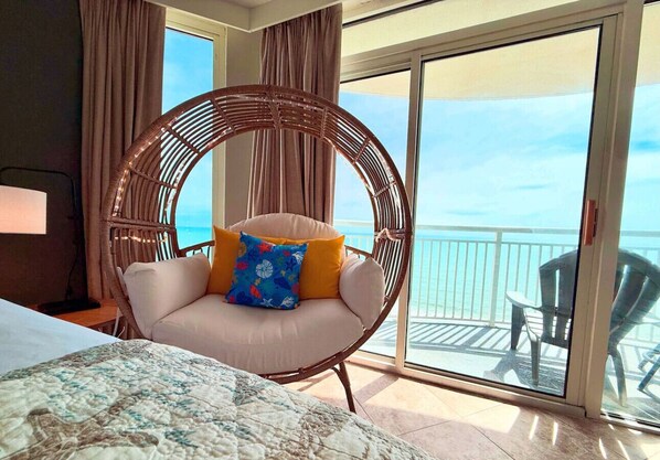 A room with a bed, a wicker chair with cushions, a view of the ocean through a glass door, and a balcony with chairs.