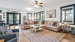 Un salon spacieux avec un grand canapé, une table basse et un tapis à motifs. Il y a deux fenêtres avec des rideaux, un ventilateur de plafond et une œuvre d’art encadrée au mur.