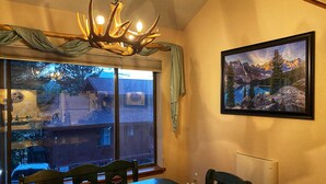 A dining area with a table and chairs, a chandelier, a large window with a view of a house, and a framed picture on the wall. - Walk To Ski Lifts! W/D, WiFi, Kitchen, Skylights (Angel Fire)