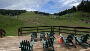 A wooden deck with green Adirondack chairs and a view of a grassy landscape with trees and a building in the distance. - Walk To Ski Lifts! W/D, WiFi, Kitchen, Skylights (Angel Fire)