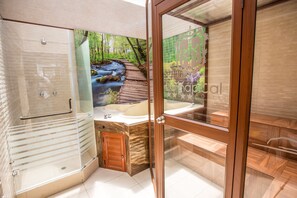 Un baño moderno con ducha de vidrio, tocador de madera y un ventanal grande con vistas panorámicas a un bosque y un arroyo.