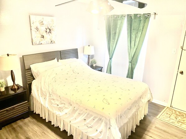 A neatly made bed with a floral bedspread, a bedside table with a lamp, and a window with green curtains.