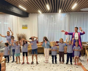 Un gruppo di bambini con camicie a righe e adulti in costume, in una stanza con pavimento piastrellato e soffitto in legno.