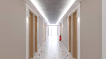 A long, narrow hallway with doors on both sides, a fire extinguisher on the right, and a bright window at the end.
