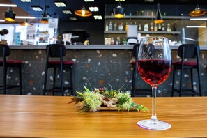 Un ambiente da bar con un bicchiere di vino rosso su un tavolo di legno, sgabelli e un bancone con bottiglie.