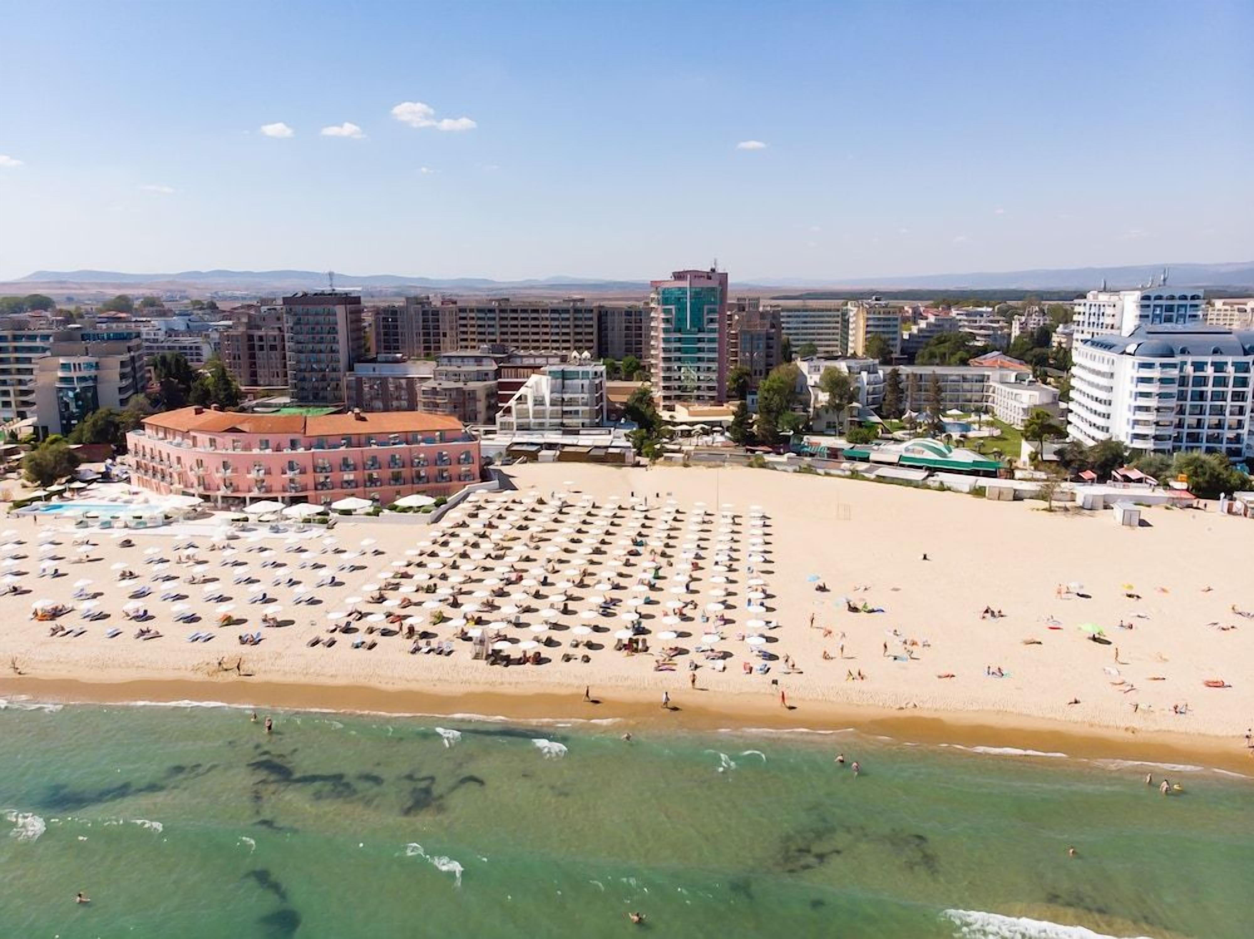 A beach with numerous umbrellas and sun loungers, a large building with a red roof, and a clear view of the ocean.