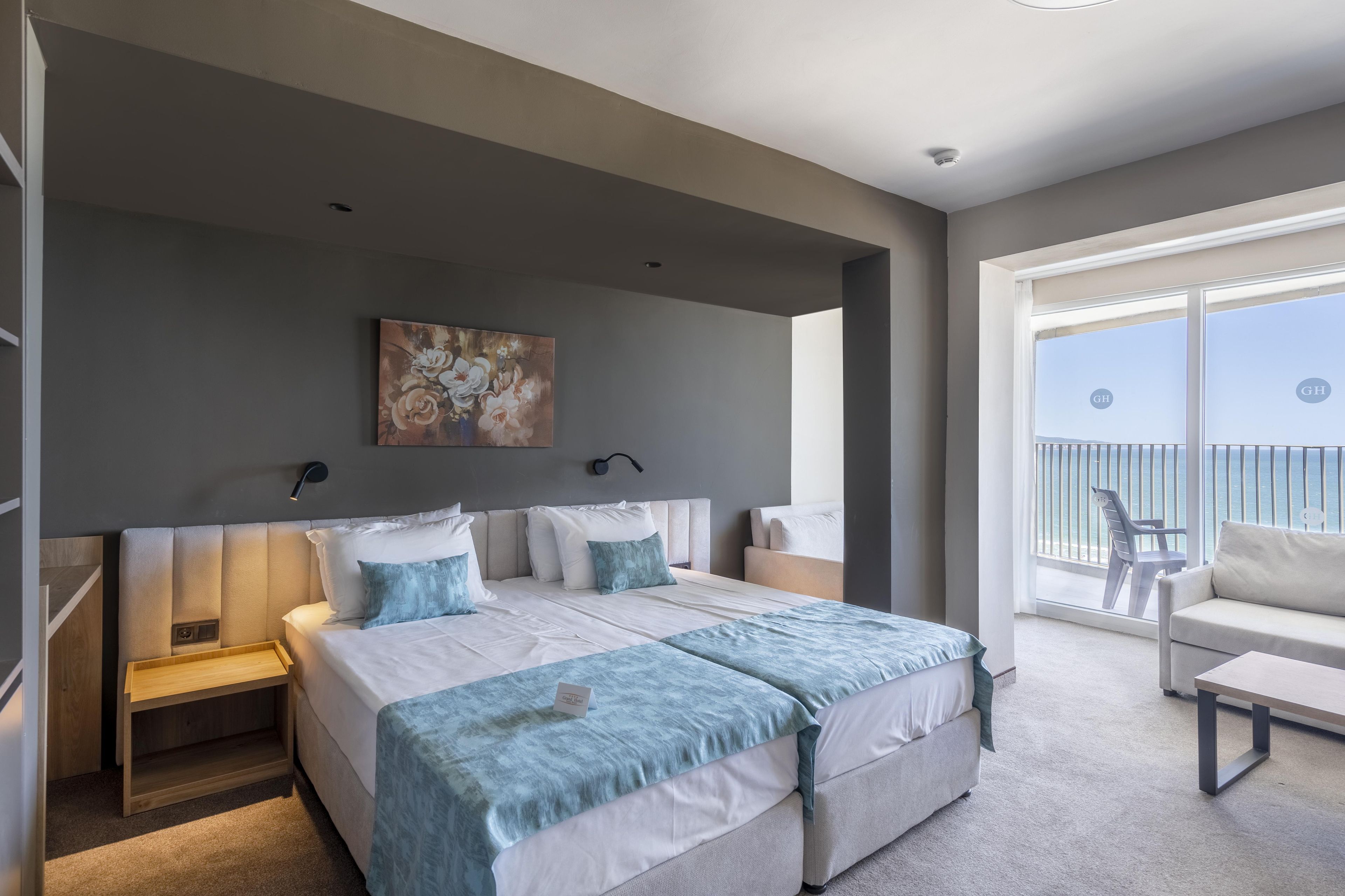 A modern bedroom with a large bed, bedside tables, a painting, and a balcony with a view.