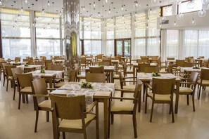 Uma sala de jantar ampla, com várias mesas e cadeiras, janelas grandes e luminárias pendentes esféricas.