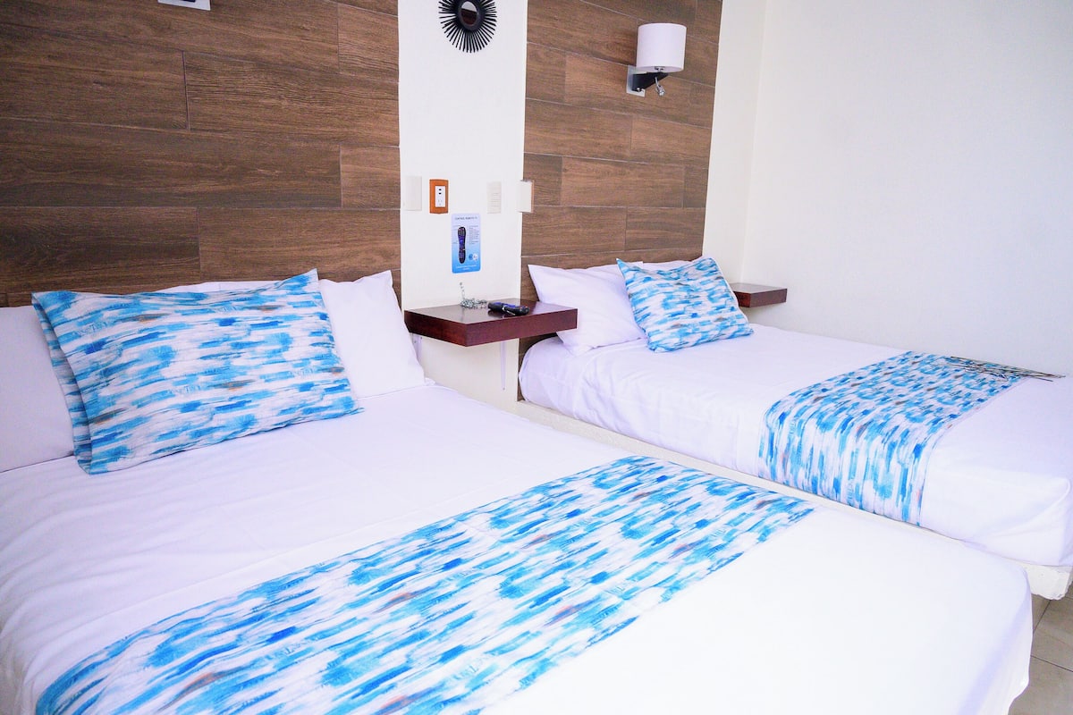 Two beds with white bedding and blue patterns, a wooden headboard, a nightstand with a lamp, and a wall-mounted light fixture.