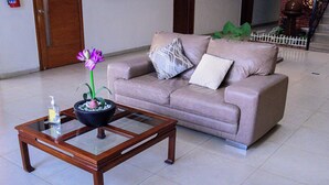 A beige sofa with a white and a patterned pillow, a glass-top coffee table with a decorative bowl and a purple flower, and a bottle of hand sanitizer. - Hotel Acuario  (Ocotlan)