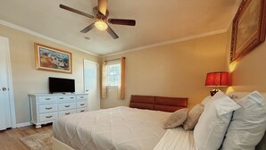 Een slaapkamer met een bed, een plafondventilator, een dressoir, een televisie, een schilderij en een raam met gordijnen.