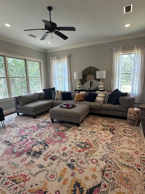 A living room with a sectional sofa, ottoman, patterned rug, ceiling fan, and large windows with curtains.