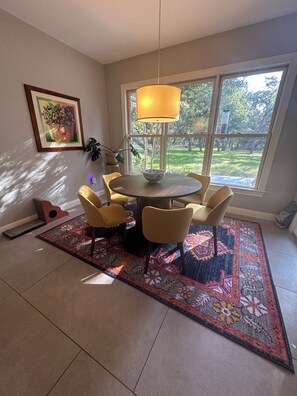 A dining area with a round table, six chairs, a hanging lamp, a framed artwork, and a view of a garden through large windows.