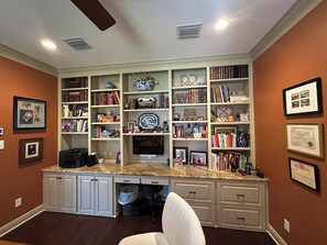 A home office with a large desk, multiple bookshelves, and framed certificates on the walls.