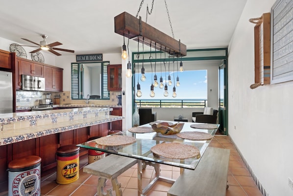Eine Küche mit Deckenventilator aus Holz, einer großen Kochinsel mit Glasplatte und Blick auf den Strand durch ein Fenster.