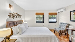 A hotel room with a large bed, a wooden headboard, a wall-mounted light fixture, two windows with wooden shutters, a striped armchair, and a small table with a lamp. - Casa D'Palma-steps to Sandy Beach! (Rincón)