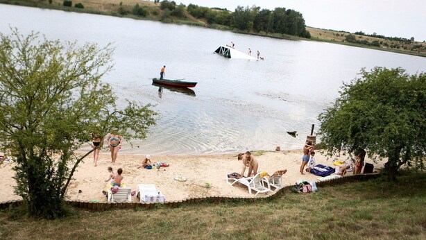 A lakeside beach with people relaxing, a small boat, and a tree-lined shore. - 2 House Between two lakes (Vilnius)