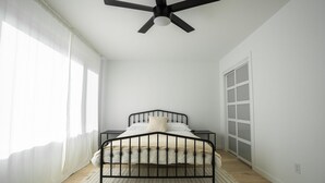 Een slaapkamer met een bed, een plafondventilator, een raam met gordijnen en een deur met geëtste glaspanelen.